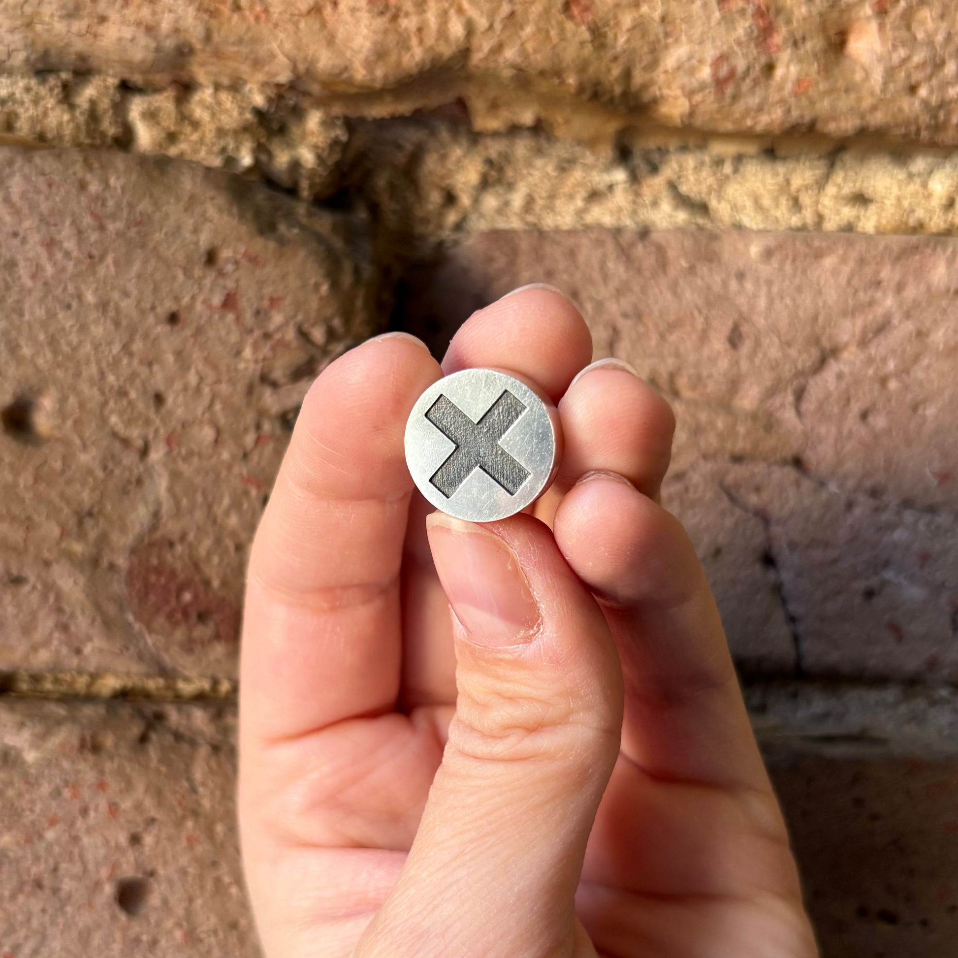 Hand holding a small metallic object with a cross symbol against a brick wall background